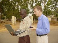 Two people measuring wireless signal strength with a laptop, standing on a dusty road in front of a gate