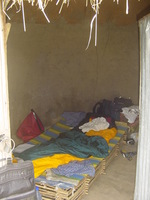 Looking through the door of a mud-plastered hut, inside is a bamboo bed with a sleeping bag on top and various bits of camping equipment scattered around
