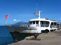 A rusty old ship sits in port.  In the distance behind it is a conical island.