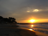 The sun hangs just above the sea at the end of a curving beach, with a glowing streak of light reflected in the water and the wet sand.