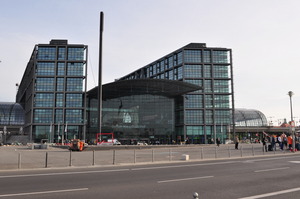 A modern building of metal and glass seen across a plaza, a sign on the front reads 'Berlin Hauptbahnhof'