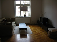 A wooden-floored room with a large sofa opposite a TV.  A large window looks out onto other apartments.