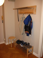 A wood and metal coat rack with a couple of coats hanging from it, attached to a large wooden board and the wall.  Below is a shoe rack.