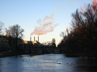 The river leads from the foreground under a small bridge.  Trees line the banks on both sides, with buildings behind the trees.  In the background three chimneys can be seen, with the outer two emitting steam.