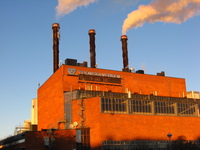 A large industrial building in red brick, glowing in the low winter sun.  Two of the three chimneys are giving off steam.
