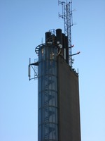 An ugly concrete chimney tower stands against the blue sky.  On the near side a spiral staircase is enclosed in a glass wall.