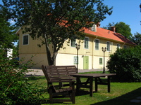 An old two-storey building in gardens.  In the foreground are a sunlit wooden bench and table.