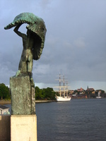On the left, in the foreground, a bronze statue of a winged man marks the start of a bridge.  To the right, in the distance, a white-painted tall ship glows in the evening sun.