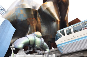 A monorail trail (with a veyr 1960's space-age design) is about to enter a tunnel through a building made of curved panels of iridescent metal.