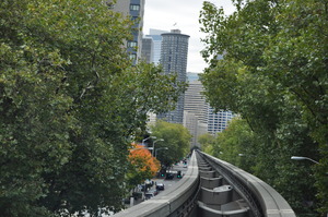 Looking ahead along monorail tracks curving and banking to the left.  Around trees on both sides of the tracks tall buildings can be seen.  Cars drive along the road below the tracks.